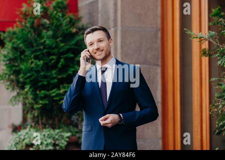 Successful businessman has work break, uses mobile phone for having talk, being always on line, dressed in formal suit, has pleasant smile on face, st Stock Photo