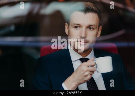 Photo of successful prosperous male entrepreneur with stubble, thinks about something, wears formal clothes, holds cup of coffee, being focused at win Stock Photo