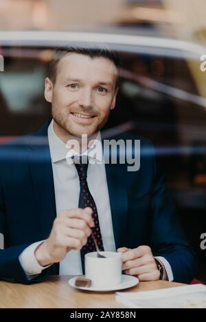 Verrtical shot of delighted handsome male professional worker in black suit, drinks aromatic beverage, looks joyfully at camera, poses in restaurant, Stock Photo