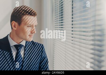 Indoor shot of pensive male economist dressed in formal outfit, looks pensively at window with jalousie conemplates about work during dinner break fee Stock Photo