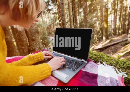 A mock-up image of a woman's hand using and typing on a laptop with a blank black desktop on a plaid grass lying in the forest. Stock Photo