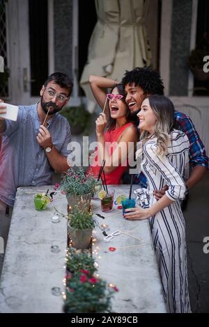 group of four adults having fun at the outdoor bar, taking selfie with funny props. Fun, party, socializing, multiethnic, couples,  concept Stock Photo