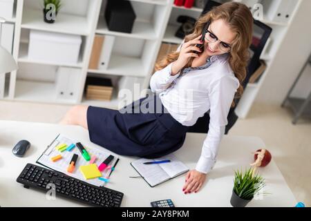 A beautiful young girl sat down on the table in the office and was talking on the phone. Before the girl there are documents. Stock Photo