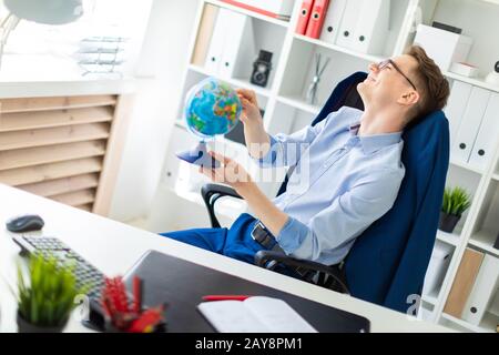 A young man sits in the office at the computer desk, closes his eyes and points a finger at the globe. Stock Photo