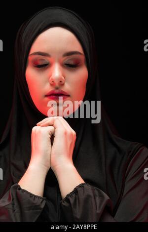Portrait of beautiful serious young muslim woman wearing black hijab with closed eyes as praying concept on black background Stock Photo