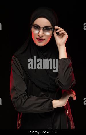 Portrait of beautiful trendy young muslim woman wearing black hijab and glasses as modern eastern fashion concept posing on blac Stock Photo
