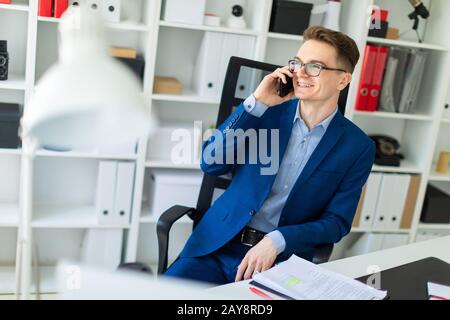 A young man is sitting at a table in the office and talking on the phone. Stock Photo