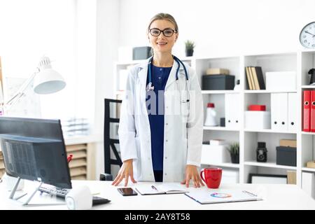 A young girl in a white coat is standing near a table in her office. A stethoscope hangs around her neck. Stock Photo