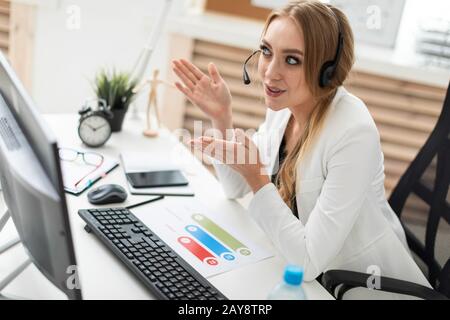 A young girl in headphones with a microphone sits at a table in the office and looks at the monitor. Stock Photo