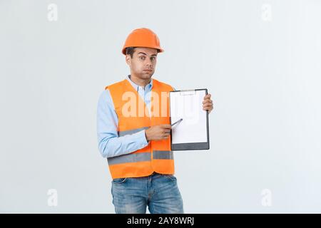 Frustrated young engineer with hardhat and reflective vest checking on mistake in document over gray background Stock Photo