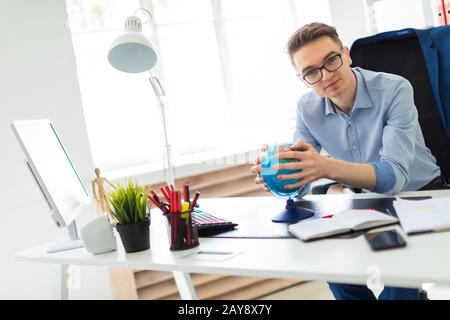 A young man sits in the office at a computer desk and holds a globe in his hands. Stock Photo