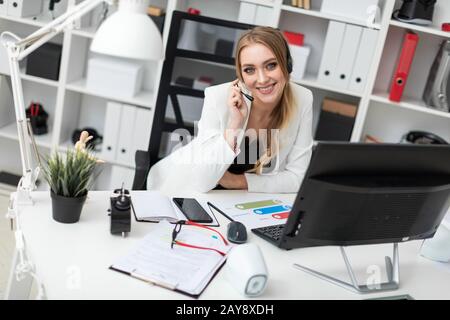 A young girl in headphones with a microphone sits at a table in the office and looks at the monitor. Stock Photo