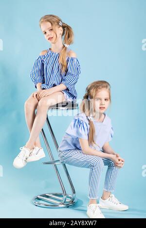 Girls twins in light blue clothes are posing near a bar stool on a blue background. Stock Photo