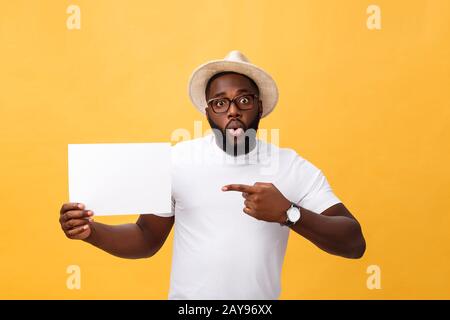 Picture of young smiling african-american man holding white blank board and pointing on it, on yellow background, copy space Stock Photo