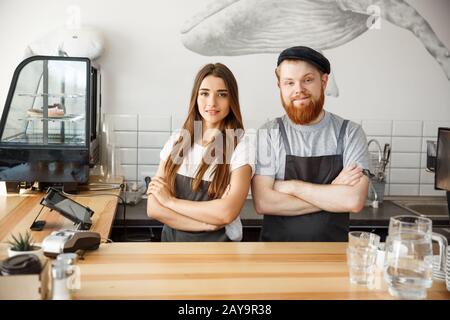 Coffee Business Concept - Positive young bearded man and beautiful attractive lady barista couple in apron looking at camera whi Stock Photo