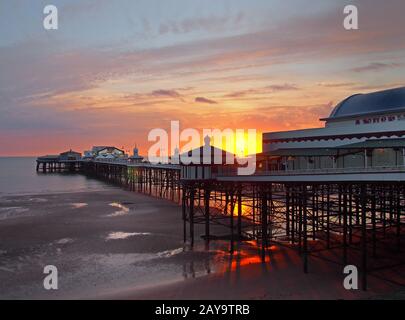 the sun setting over the historic north pier in blackpool with glowing light reflected on the beach Stock Photo