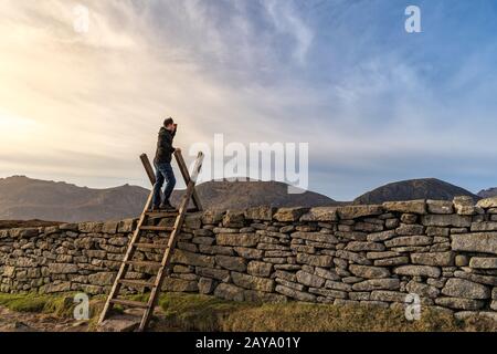 Man standing on the ladder on the stone wall in the mountains with sunset in background Stock Photo
