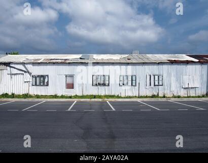 dilapidated abandoned white tin sheet corrugated buildings with empty car park blue sky and clouds Stock Photo