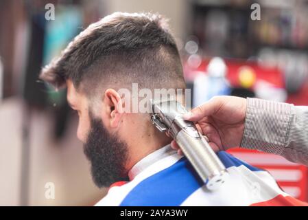 Handsome bearded man, getting haircut by barber, with electric trimmer at barbershop. Stock Photo
