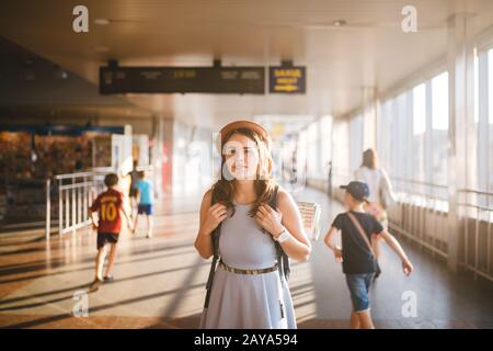 Theme tourism travel. Young beautiful Caucasian woman in dress and hat with backpack tourist mat in terminal station in a long w Stock Photo