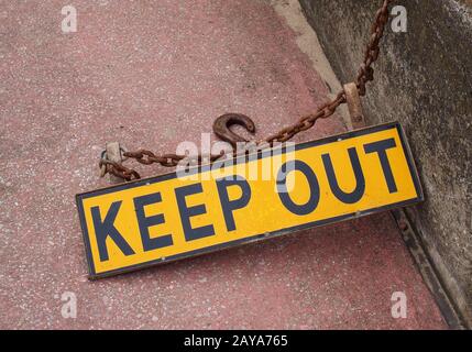 a metal fluorescent yellow keep out sign on a concrete floor with rusty chain attaching it to a wall with an iron hook Stock Photo