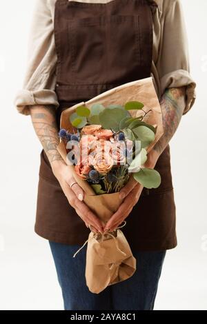 Woman in a brown apron is holding summer wonderful fresh roses, eryngium and green leaves as a greeting bunch on a light backgro Stock Photo