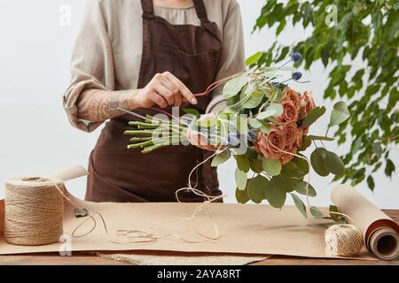 Female florist is decorating beautiful bouquet from fresh natural roses step by step at the table with paper and rope on it. Mot Stock Photo