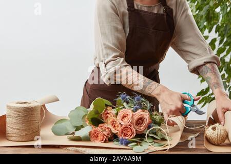Female florist is cutting paper for creating beautiful bouquet on a background of light wall. Place for text. Process of making Stock Photo