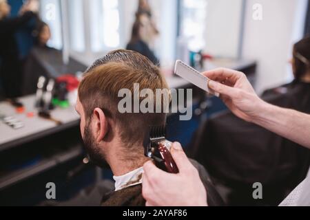 Close up shot of man getting trendy haircut at barber shop. Male hairstylist serving client, making haircut using machine and co Stock Photo