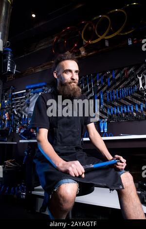 Portrait of small business owner of young man with beard. Guy bicycle mechanic workshop worker sitting with tool in his hand in Stock Photo
