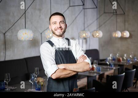 A young, smiling waiter in a restaurant, standing next to the tables with a glass of wine. He wears an apron, looks confidently, Stock Photo