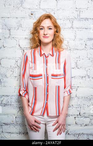 A beautiful young woman student with red curly hair and freckles is leaning against a brick wall of gray color. Dressed in a red Stock Photo