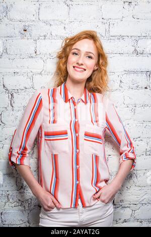 A beautiful young woman student with red curly hair and freckles is leaning against a brick wall of gray color. Dressed in a red Stock Photo