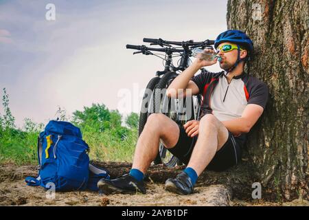 Caucasian male cyclist active lifestyle resting under tree after exercising bicycle drinking water from bottle. Sportsman in hel Stock Photo