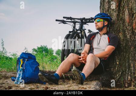 male mountain biker resting on bike ride, sitting on ground under a tree with his mountain bike, stands next to him, enjoying th Stock Photo