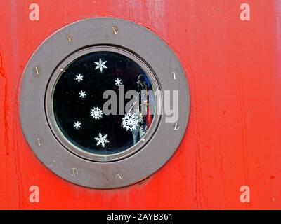 a close up of a round metal porthole on a red boat with snowflakes and glitter on the window and shiny charms hanging inside Stock Photo