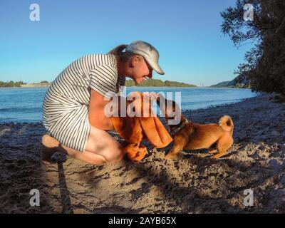 Woman playing with her small mixed breed dog at the river beach with a towel. Dog, lifestyle and sum Stock Photo