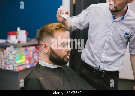Barber Making Haircut Bearded Man In Barbershop. Professional stylist cutting client hair in salon. Barber using scissors and co Stock Photo
