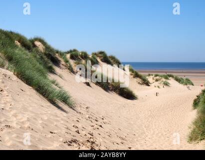 a path through tall grass covered sand dunes leading to a beach and bright blue sea in summer sunlight on the sefton coast in me Stock Photo