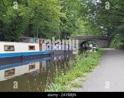 narrow boats and barges moored on the rochdale canal in hebden bridge bext to an old stone footbridge surrounded by green summer Stock Photo