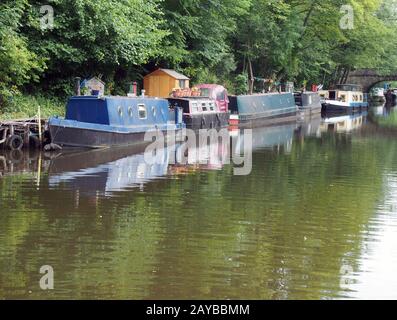 narrow boats and barges moored on the rochdale canal in hebden bridge surrounded by green summer trees and a stone footbridge Stock Photo