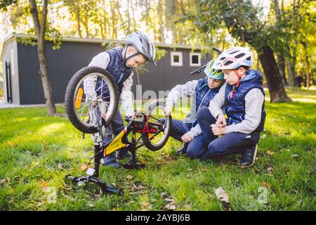Teamwork. big family three Caucasian children brothers are learning to repair a bicycle, use tools in helmets same clothes, agai Stock Photo