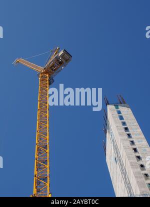 a yellow tower crane working on a construction site with a tall concrete building against a bright blue sky Stock Photo