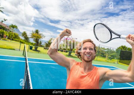 Tennis player man winning cheering celebrating victory. Winner man happy in celebration of success and win. Fit male athlete on tennis court outdoors holding tennis racket in triumph by the net. Stock Photo