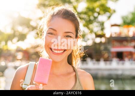 Happy summer treat Asian girl eating strawberry ice cream stick outside in sun flare sunset. Smiling Chinese woman smiling enjoying city park lifestyle. Stock Photo