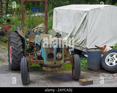 an old rusting abandoned tractor next to a dirty tarpaulin covered trailer and junk in a farmyard Stock Photo