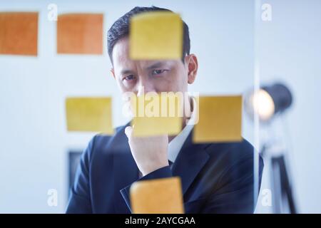 Asian pensive businessman  looking at glass wall with sticker notes Stock Photo
