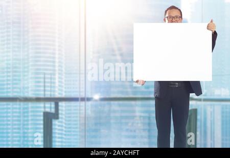 Businessman standing and holding white empty board in front of windows office overlooking the city at sunrise . Stock Photo