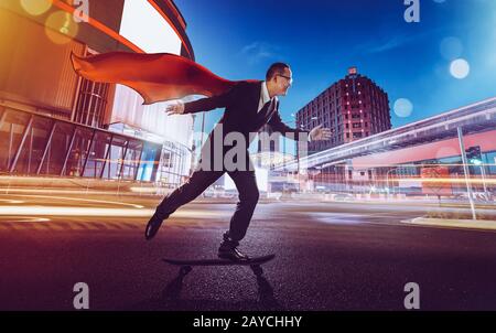 Young confident super businessman in red cape and  black suit on a skateboard going fast . night scene with modern city . Stock Photo