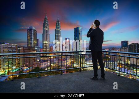 Young businessman standing on a balcony  using smartphone with beautiful city skyline background . Night scene . Stock Photo